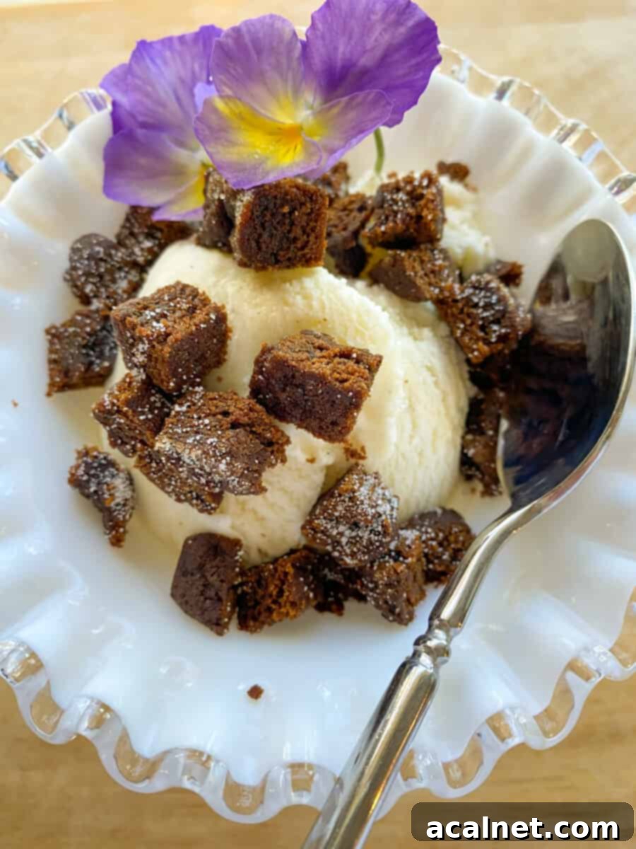 A scoop of vanilla ice cream crowned with diced molasses snap pieces and garnished with a beautiful pansy flower and a spoon.