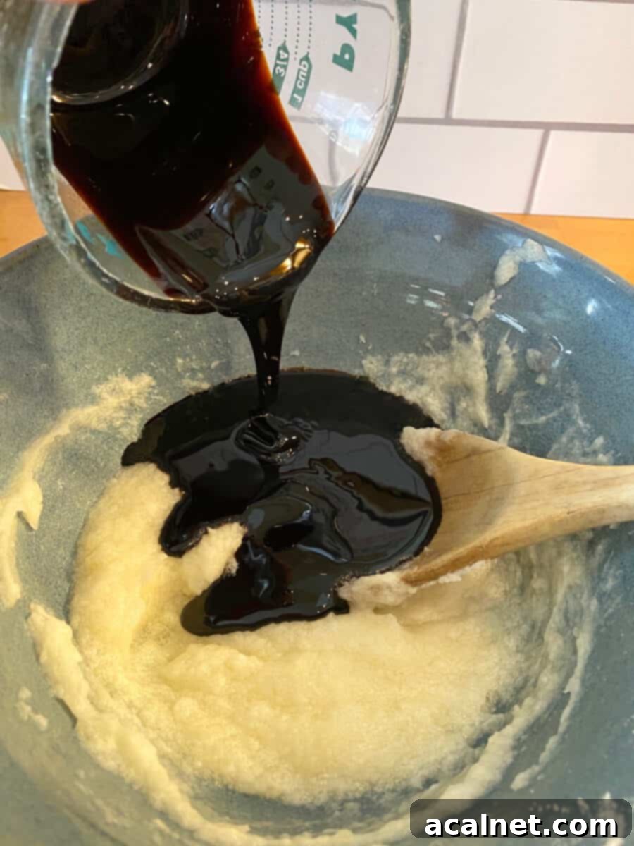 Thick, dark molasses pouring generously into a mixing bowl filled with creamed ingredients, creating a rich swirl.