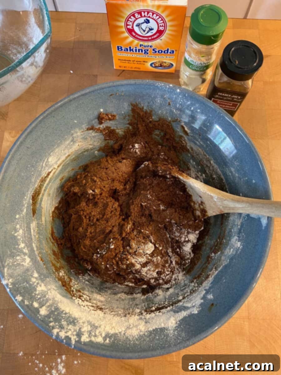 A mixing bowl contains the thick, rich molasses snap dough, with a sturdy wooden spoon resting within it and spice containers in the background.