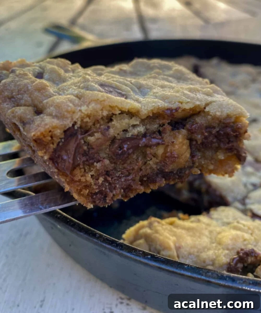 Chocolate chip bar cookie on a spatula, lifted out of a cast iron skillet on an outdoor table. Each bar cookie is brimming with delicious melted chocolate chips and crunchy walnuts.