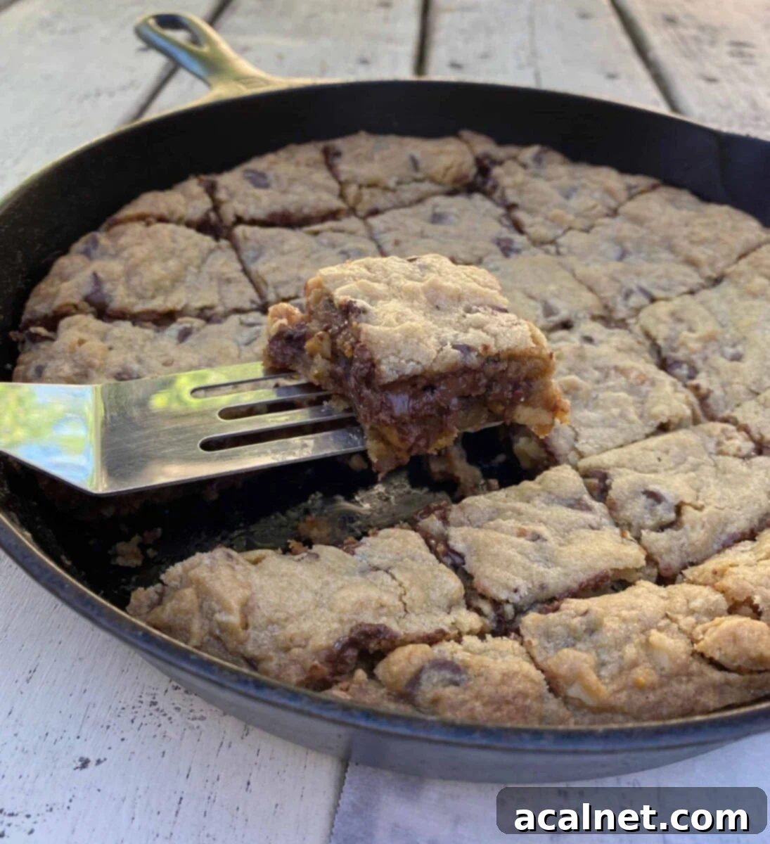A delicious chocolate chip bar cookie is lifted on a spatula from a cast iron skillet, showcasing its chewy texture and melted chocolate.