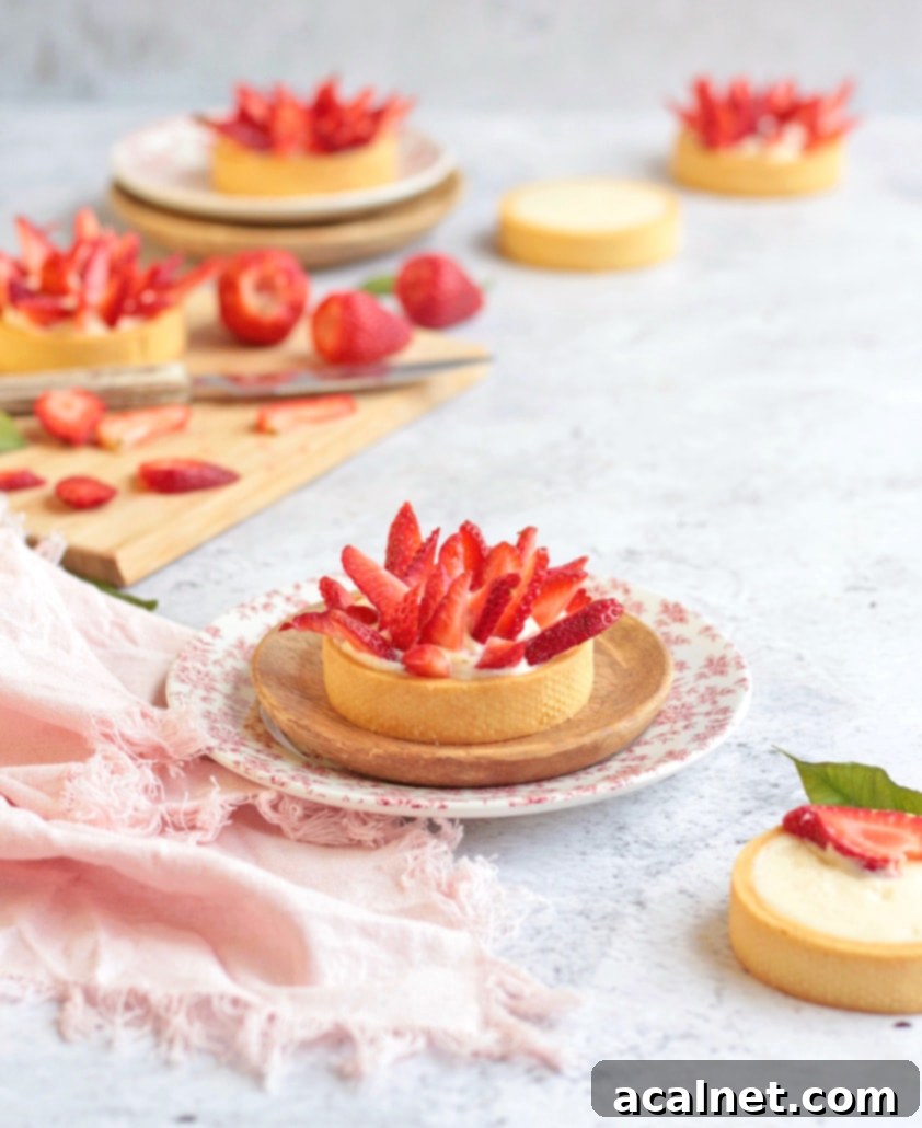 A small, perfectly assembled strawberry tartlet on a plate with a pink napkin, with a wooden cutting board of fresh strawberries in the background.