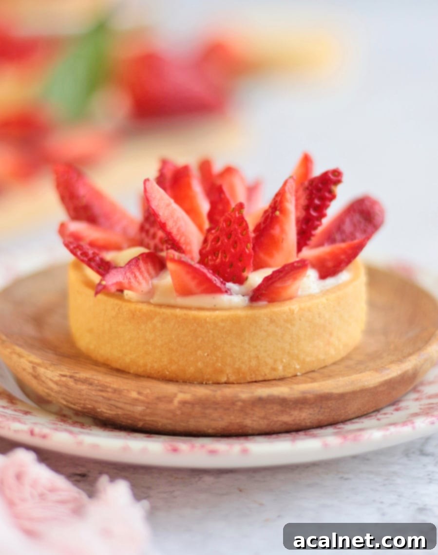 Close-up of a single beautifully finished strawberry custard tartlet resting on a small wooden plate.