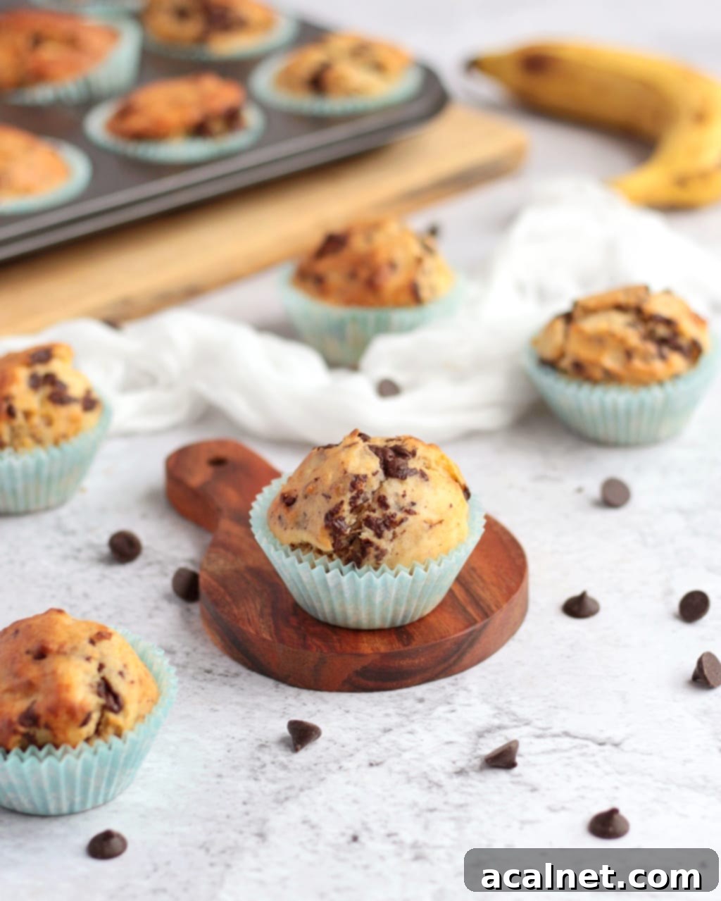 One freshly baked banana muffin on a small wooden board, surrounded by more golden muffins and scattered dark chocolate chips.