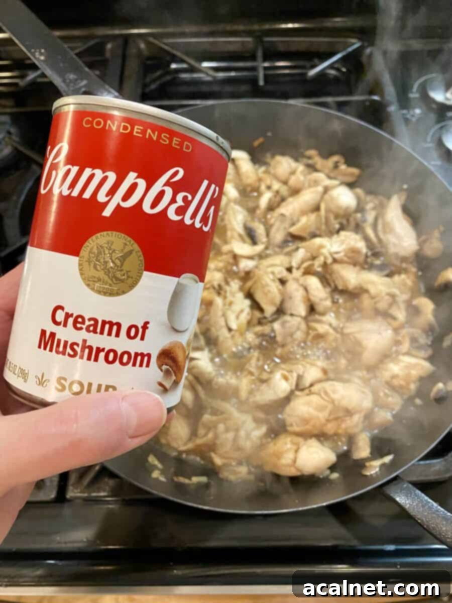 Creamy Chicken and Mushroom Stroganoff 5 A can of cream of mushroom soup being held over a pan of chicken and mushrooms to be added to the sauce.