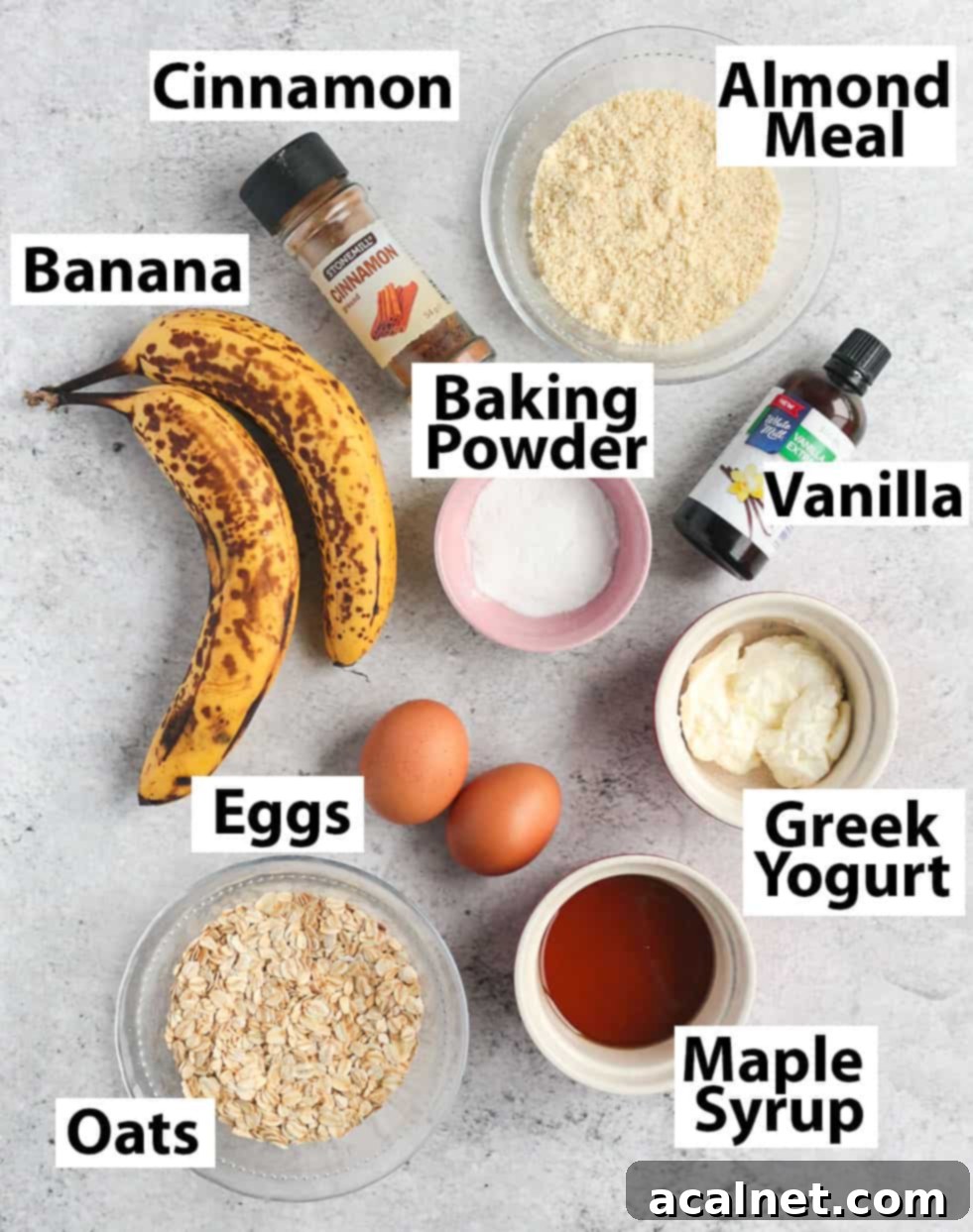 A selection of ingredients for making oatmeal banana muffins laid out on a grey surface, including oats, bananas, eggs, and syrup.