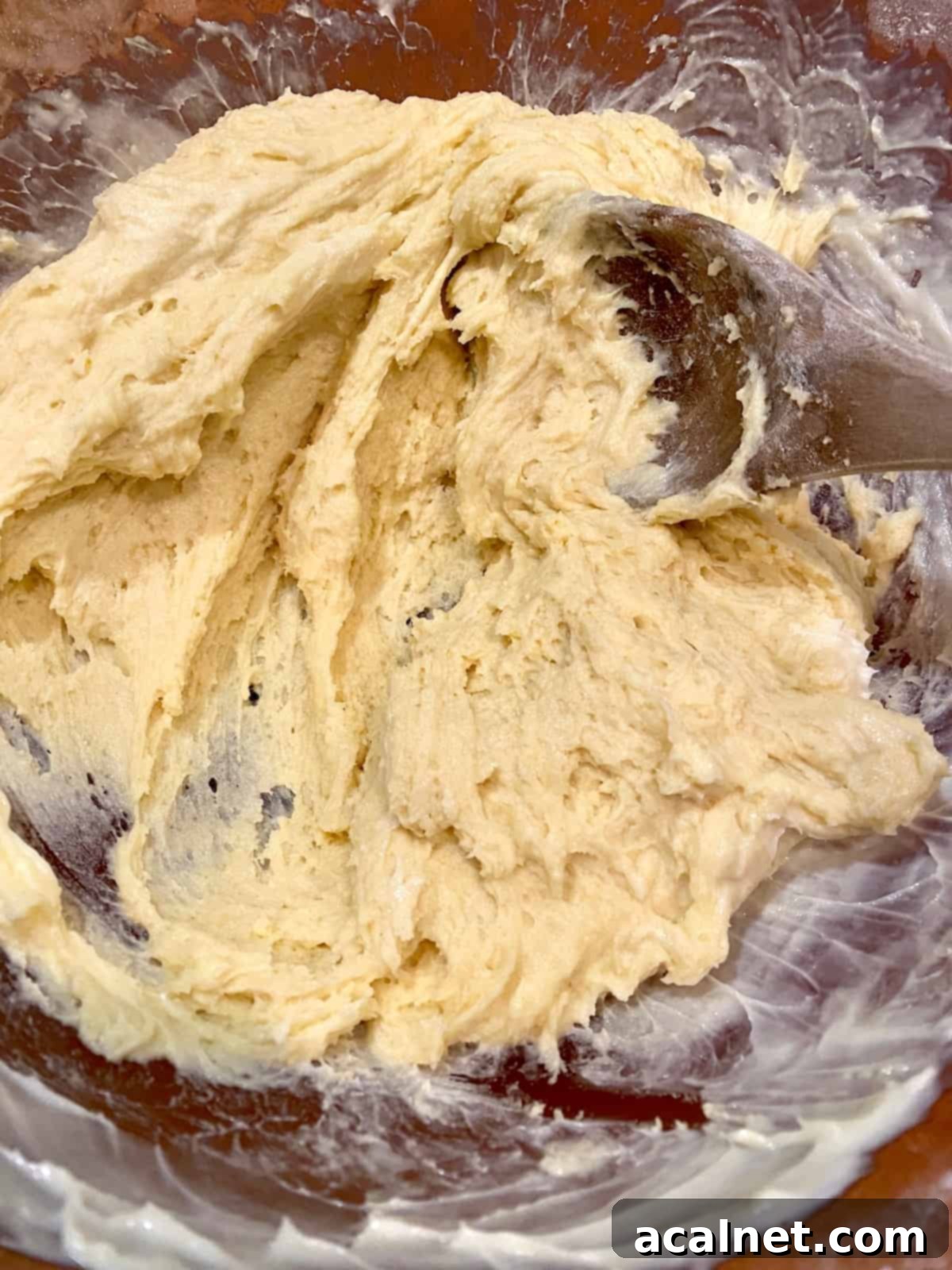 A wooden spoon stirring the thick, sturdy muffin dough in a bowl, showing flecks of flour still being combined with the creamy batter, indicating the correct consistency.