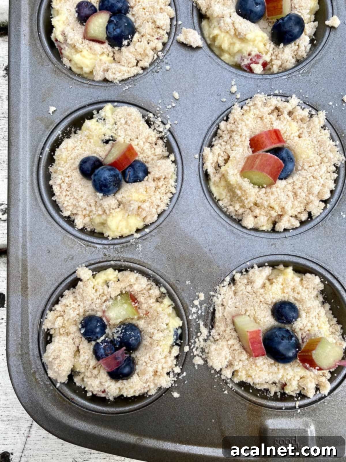 A muffin pan filled with unbaked blueberry rhubarb muffins, generously topped with fruit and batter, awaiting their streusel and baking process.