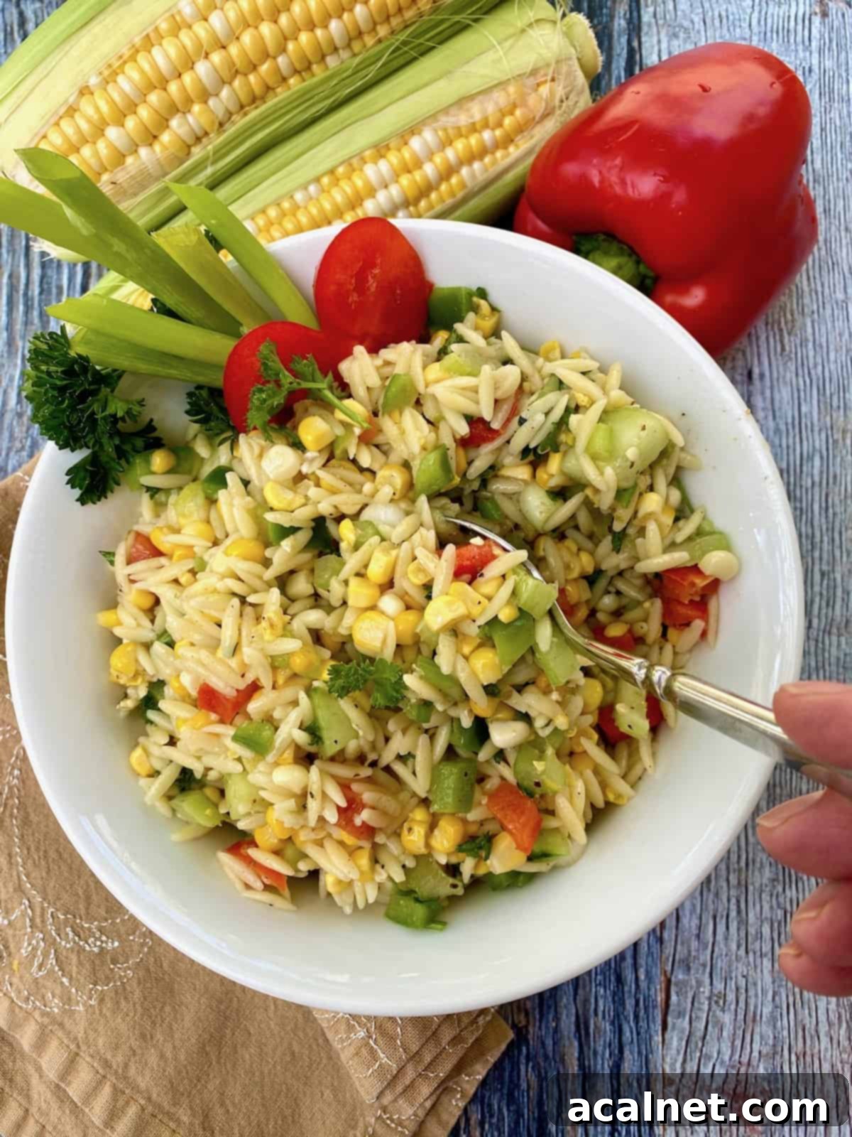 A spoon serving a portion of sweet corn pasta salad from a bowl with several ears of corn and a red pepper in the background.