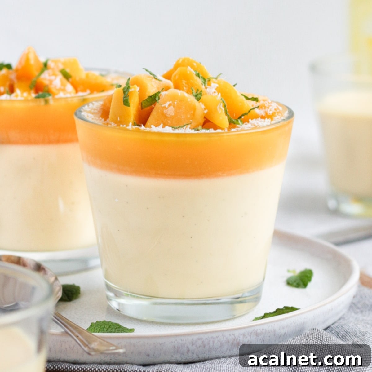 Close up on a panna cotta in a small glass cup placed over a white plate with fresh mango and mint