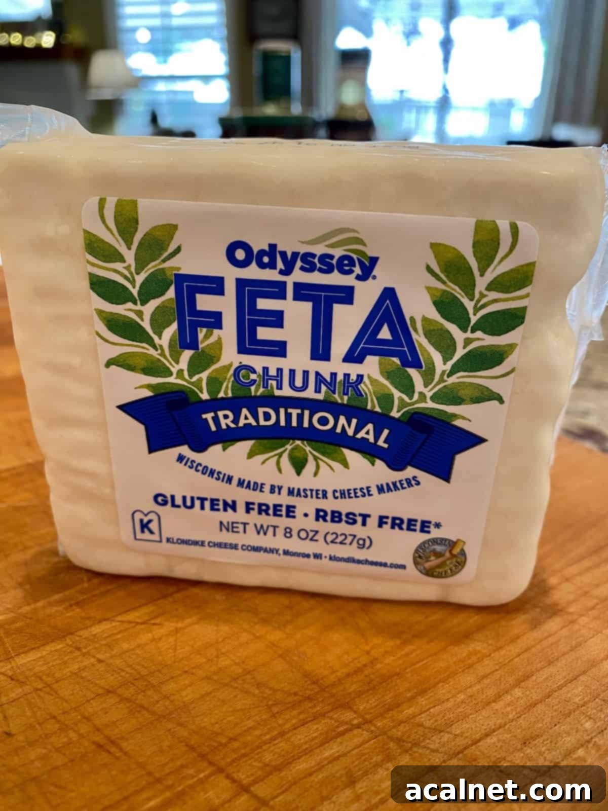 A single, unwrapped block of feta cheese rests on a wooden cutting board, ready to be prepared.