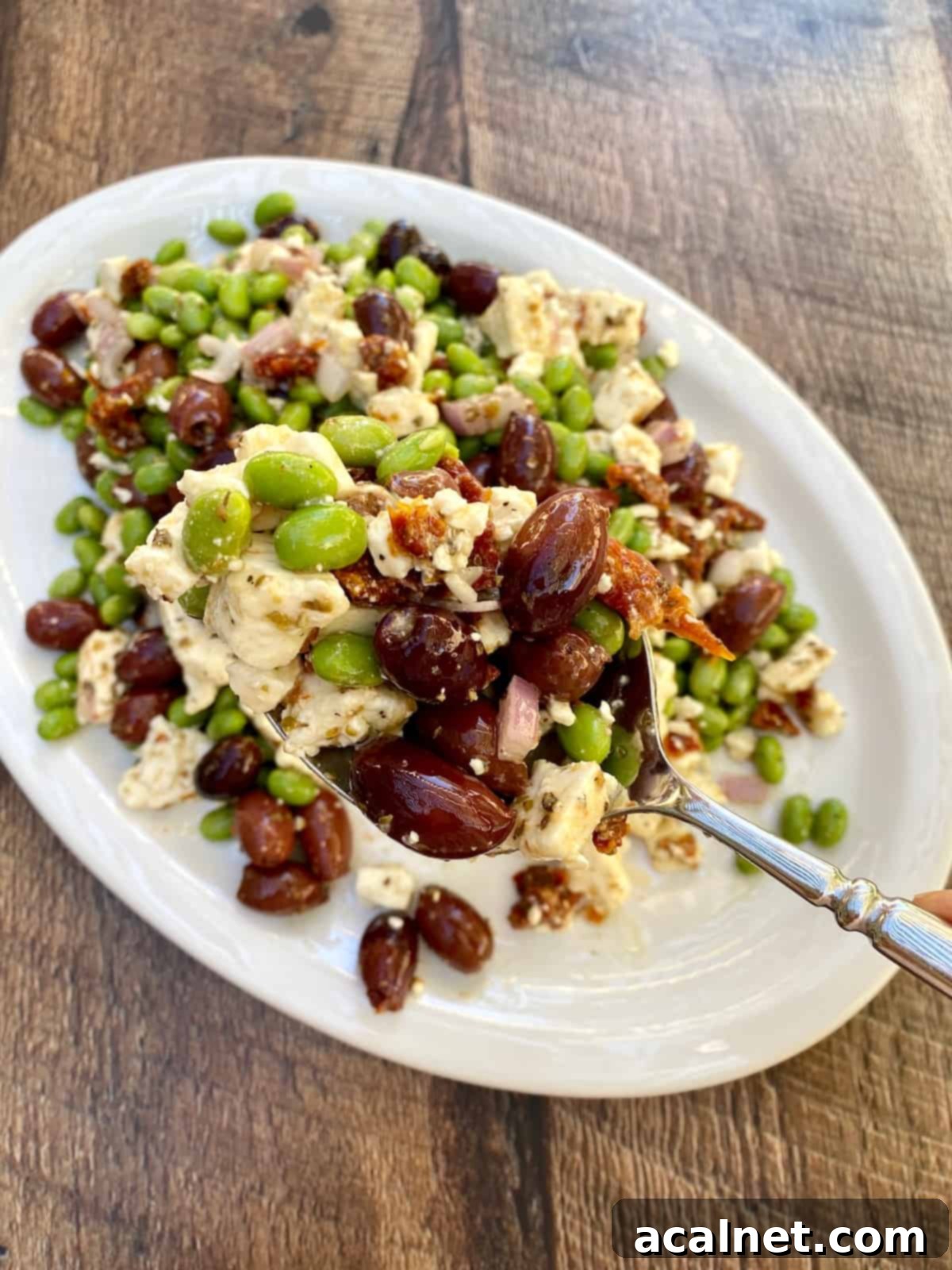 A vibrant Mediterranean Edamame Salad is beautifully displayed on a white plate, with a large serving spoon lifting a portion, ready to be enjoyed.