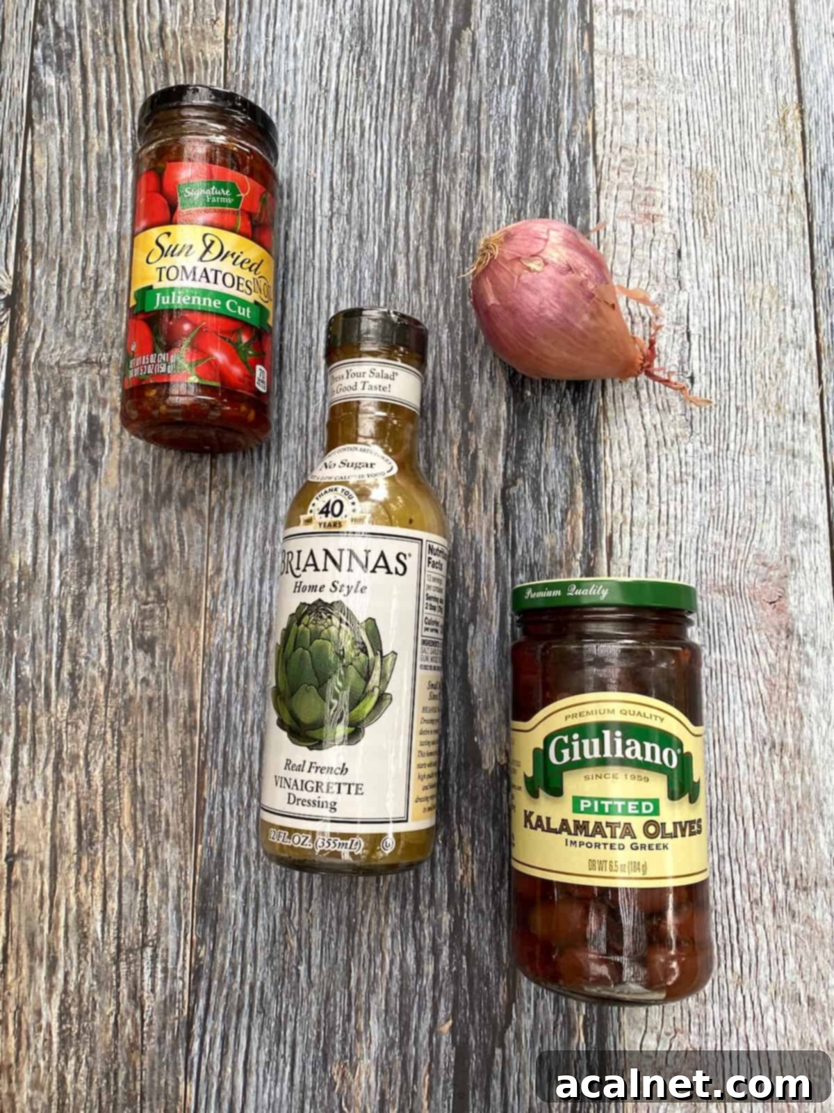 Various ingredients for edamame salad, including bottles of sun-dried tomatoes, a jar of olives, salad dressing, and a shallot, arranged on a rustic wooden plank.