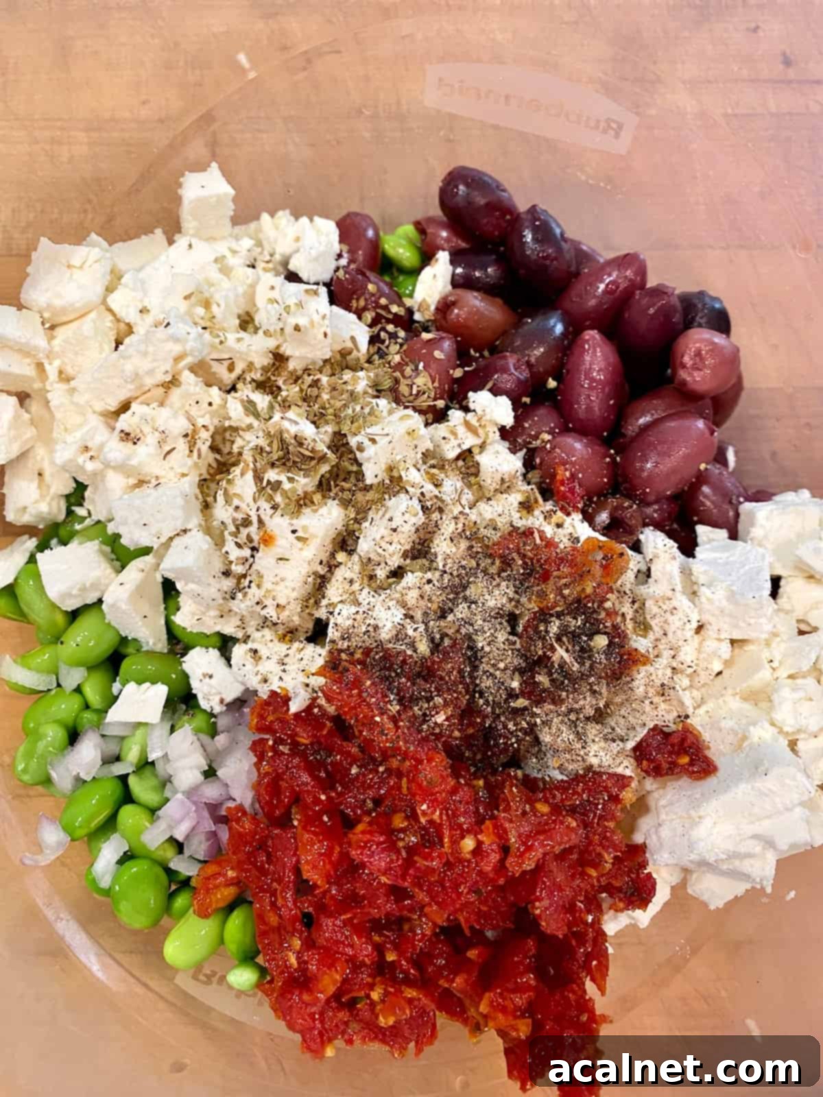 All the colorful ingredients for the edamame salad, including green edamame, white feta, and red sun-dried tomatoes, are gathered in a large mixing bowl, ready to be tossed.