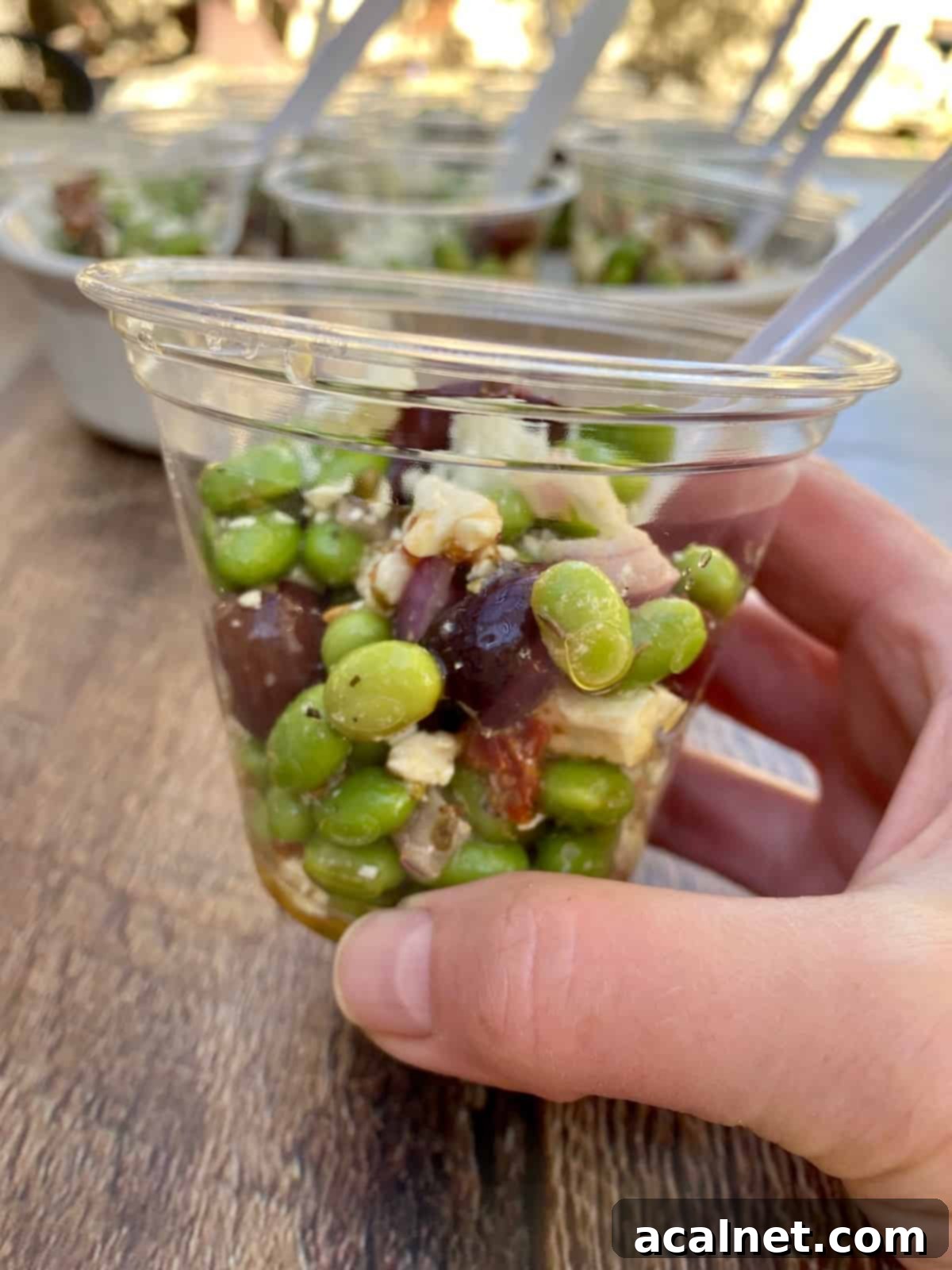 A single serving of Mediterranean Edamame Salad is neatly presented in a clear, small plastic cup, ideal for individual portions at events.