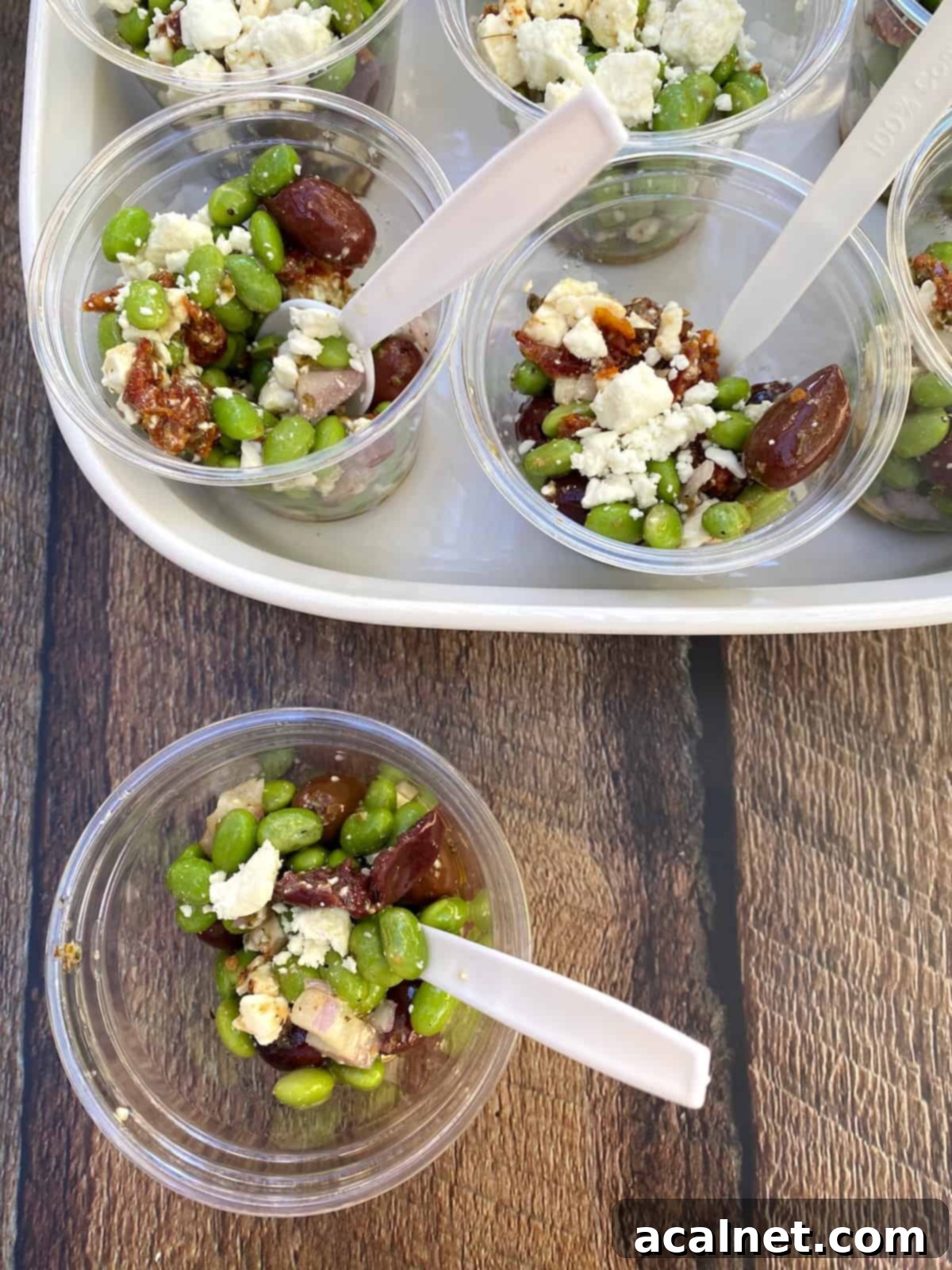 A serving dish filled with multiple small clear cups, each containing a portion of edamame salad with a spoon, and one cup artfully placed to the side.