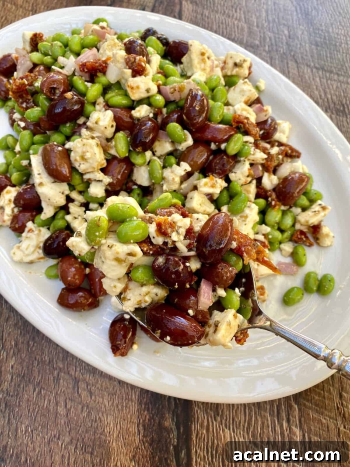 A large white plate showcasing a generous spread of Mediterranean Edamame Salad, with a serving spoon poised to scoop a portion, highlighting its vibrant colors and textures.