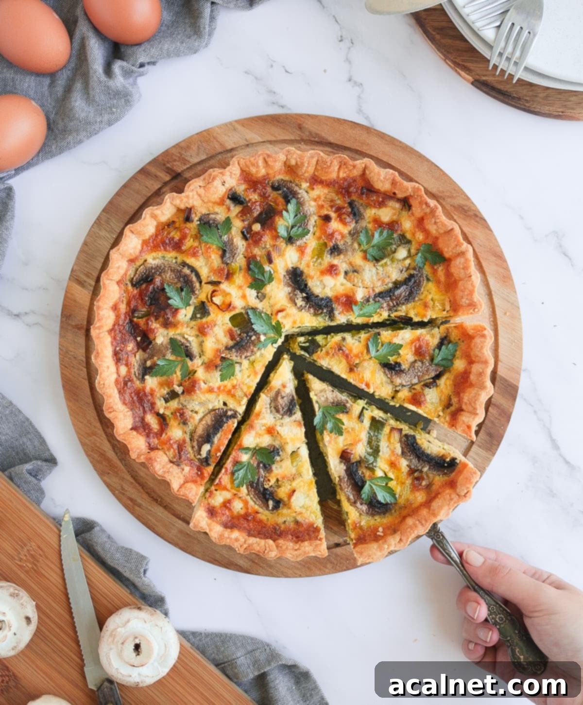 Quiche from above on a round wooden board with 3 slices cut and a hand picking up one slice, showing the inviting texture.