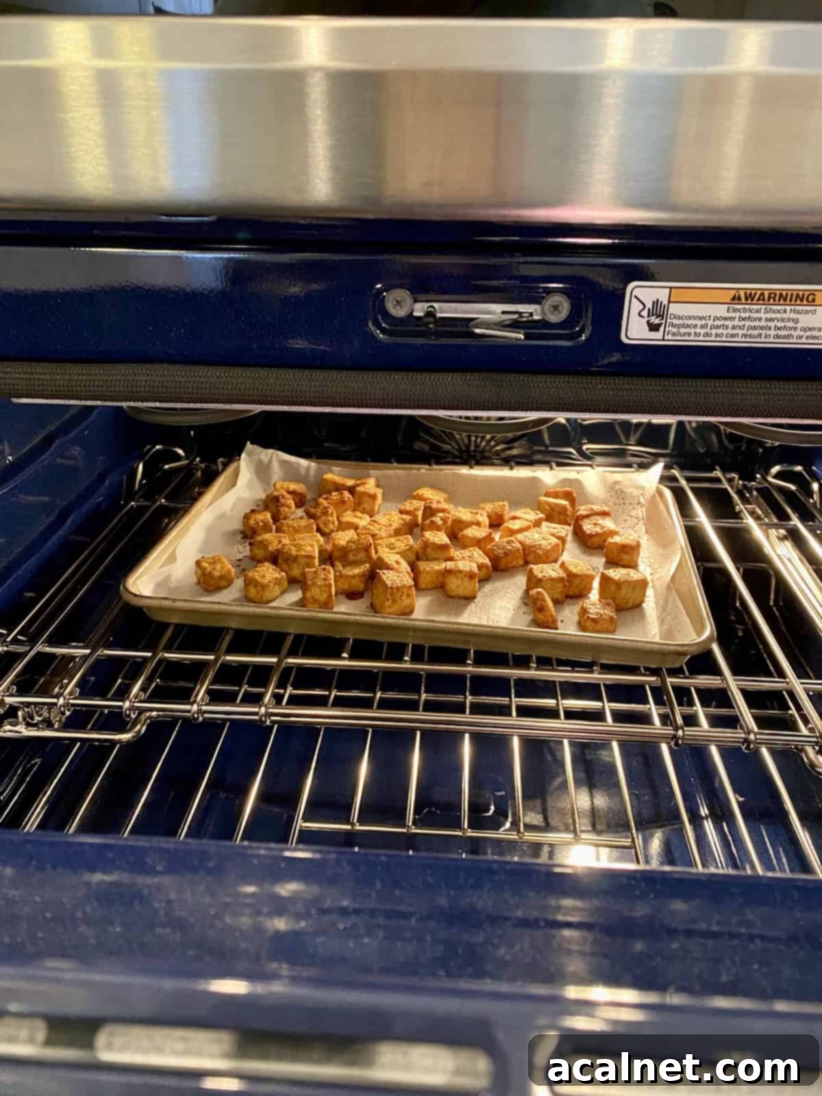 A baking sheet filled with evenly spaced, diced tofu chunks on parchment paper, inside an oven, baking to crispy perfection.