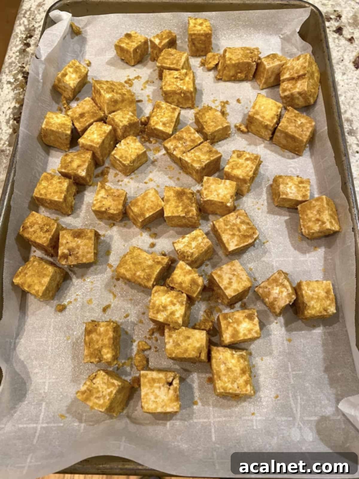 Crispy, diced tofu pieces neatly arranged on parchment paper on a baking sheet, golden brown and ready to be sauced.