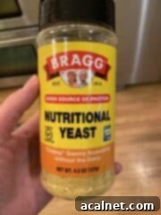 A hand holding a clear plastic jar of nutritional yeast flakes.