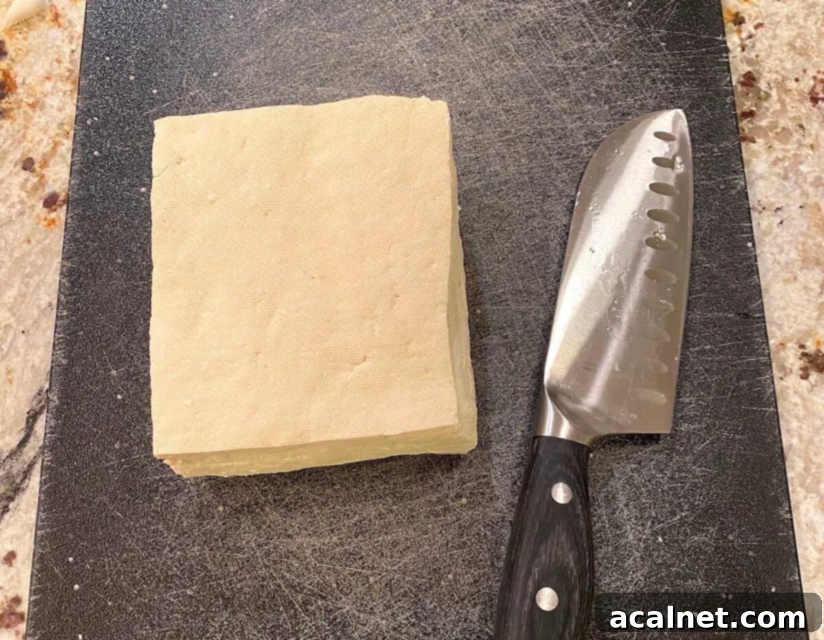 A block of extra firm tofu on a wooden cutting board with a knife beside it, ready for pressing.