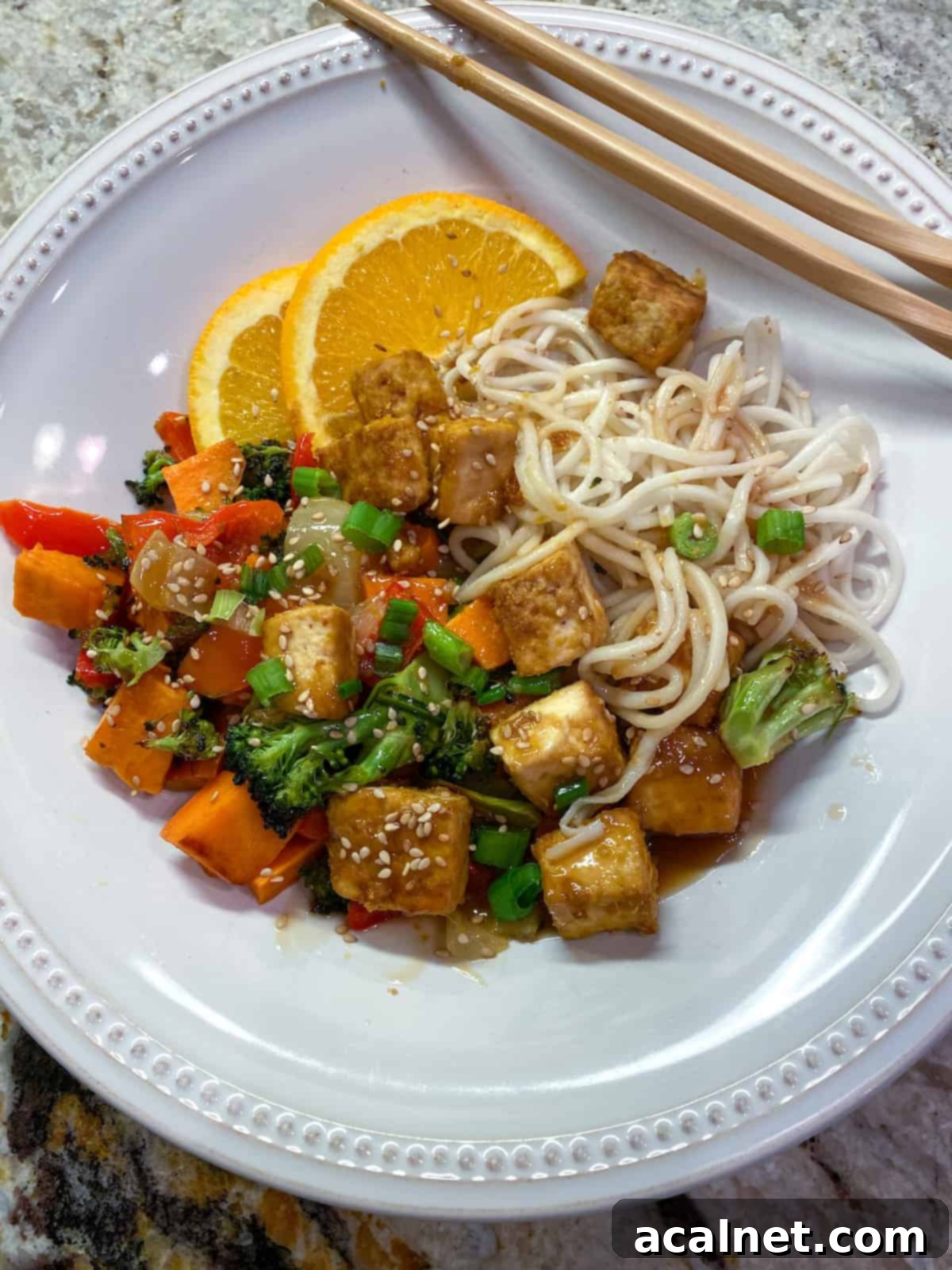 A close-up of baked orange tofu with steamed vegetables and noodles, drizzled with sauce and garnished.