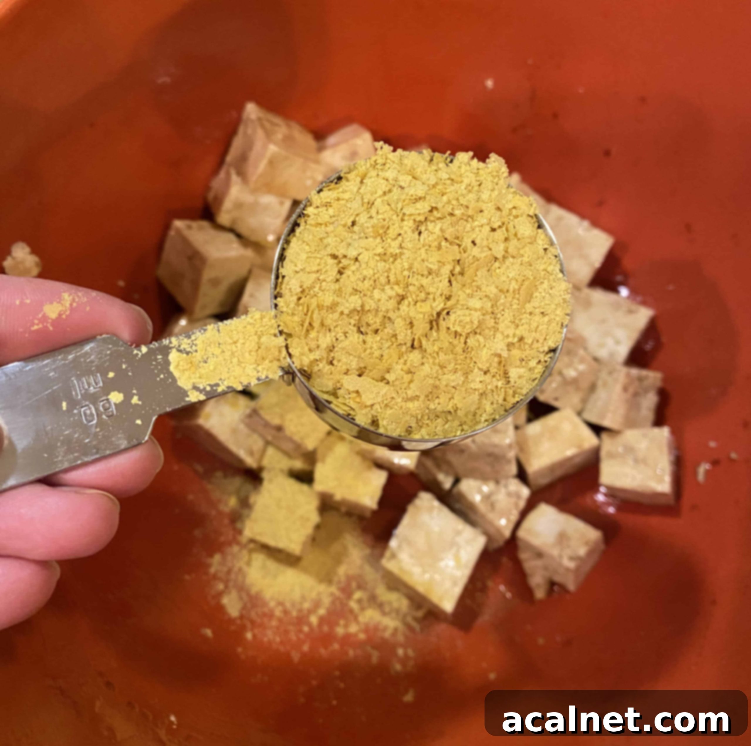 A hand sprinkling nutritional yeast over a bowl of diced tofu coated in soy sauce and olive oil.
