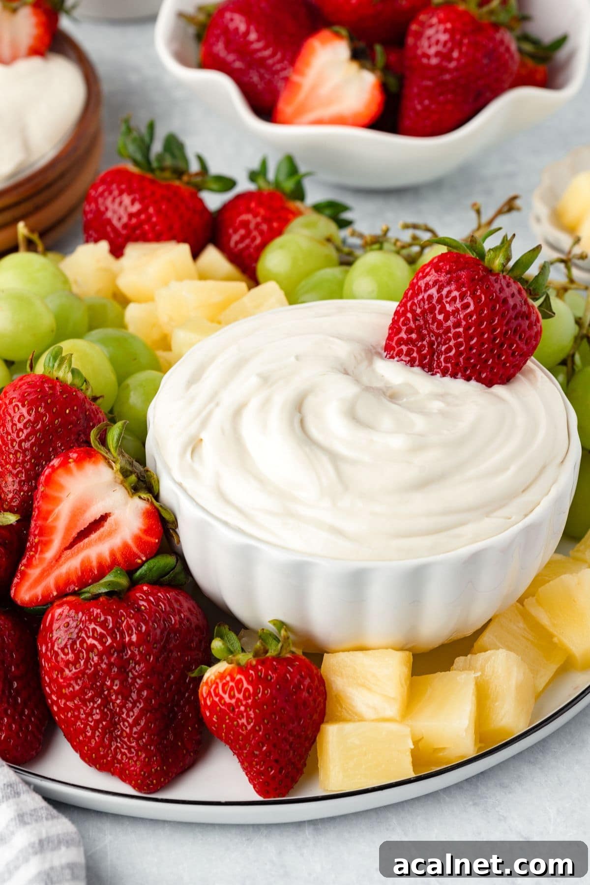 A white bowl of Marshmallow Fruit Dip sits on a platter, creamy and inviting, topped with a whole strawberry and surrounded by fresh strawberries, green grapes, and pineapple chunks.