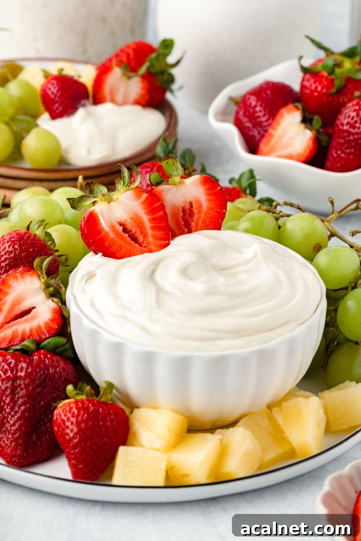 A white bowl filled with Marshmallow Fruit Dip is surrounded by fresh strawberries, green grapes, and pineapple chunks on a platter. More strawberries and grapes are in dishes in the background.
