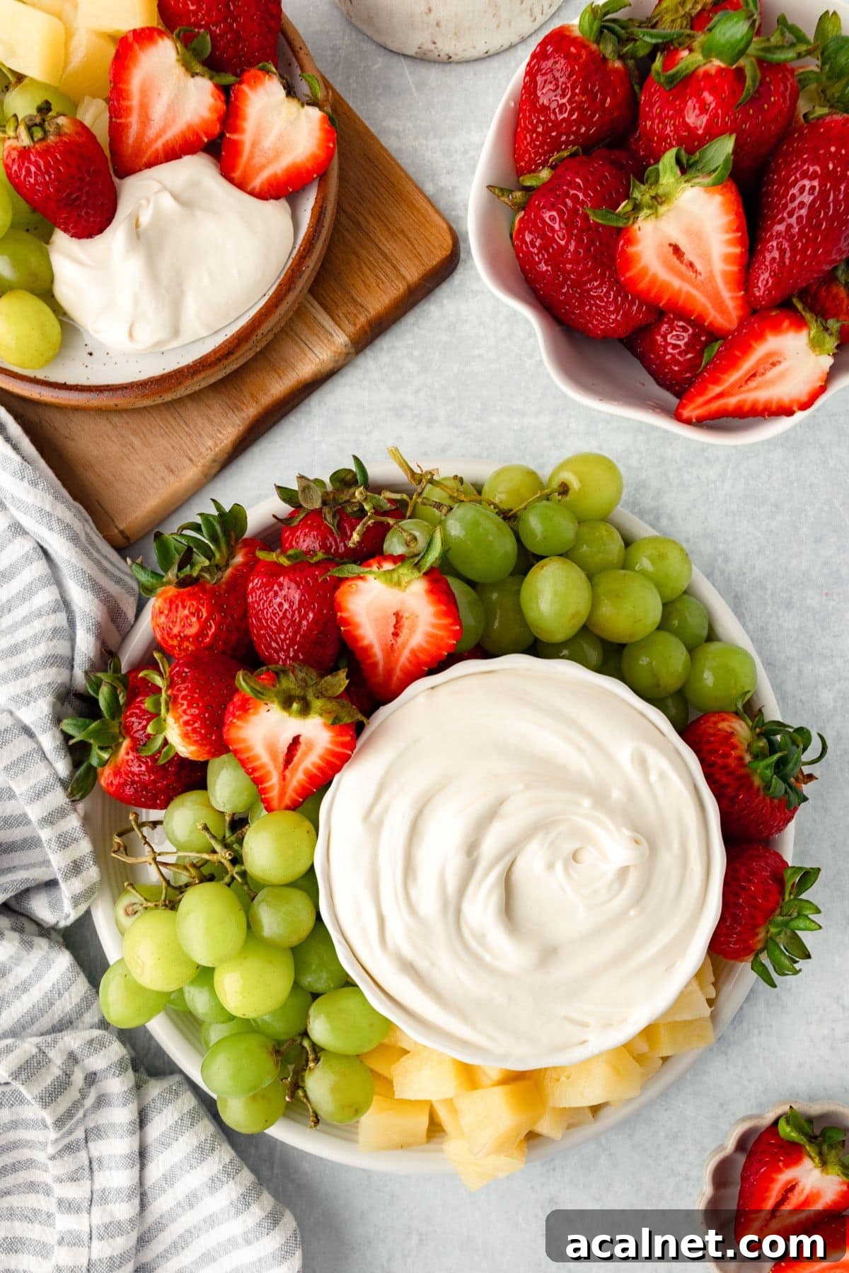 A platter with fresh strawberries, green grapes, and pineapple chunks surrounds a bowl of creamy Marshmallow Fruit Dip. Nearby, a smaller plate holds more fruit and dip, while a bowl is filled with whole strawberries on a striped cloth.