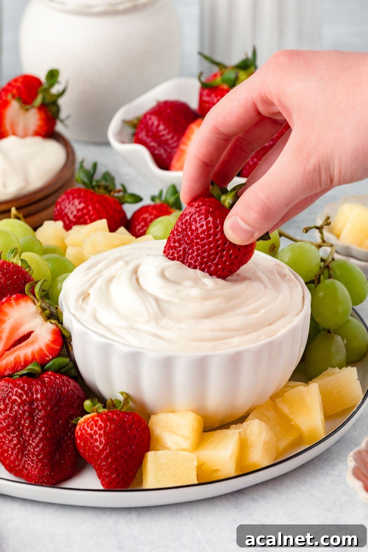 A hand dips a fresh strawberry into a bowl of Marshmallow Fruit Dip, surrounded by grapes, pineapple chunks, and more strawberries on a white platter.