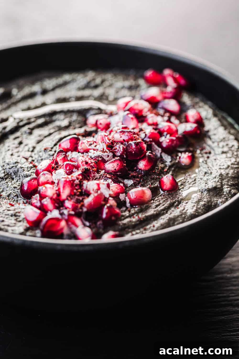 black hummus topped with pomegranate seeds in a black bowl, side angle