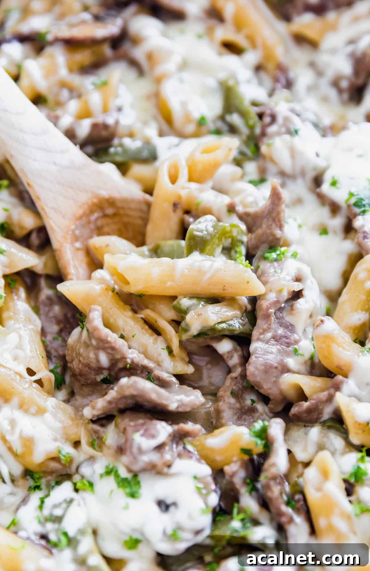 A close up of a wooden spoon in cheesy Philly pasta with beef and peppers and mushrooms.