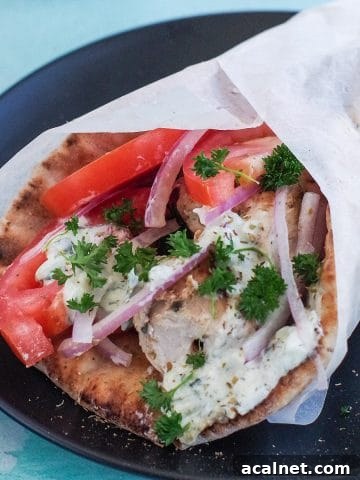 Greek chicken souvlaki wrapped in pita with tomatoes, red onion, tzatziki and parsley, wrapped in paper on black plate
