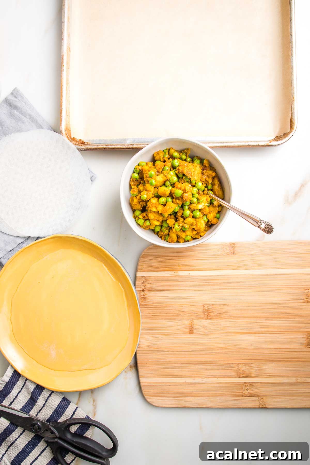 Everything ready to begin making samosas: rimmed baking tray lined with parchment, stack of rice paper wrappers, rimmed dish filled with water, samosa filling, small wooden board, kitchen scissors and a dish towel.