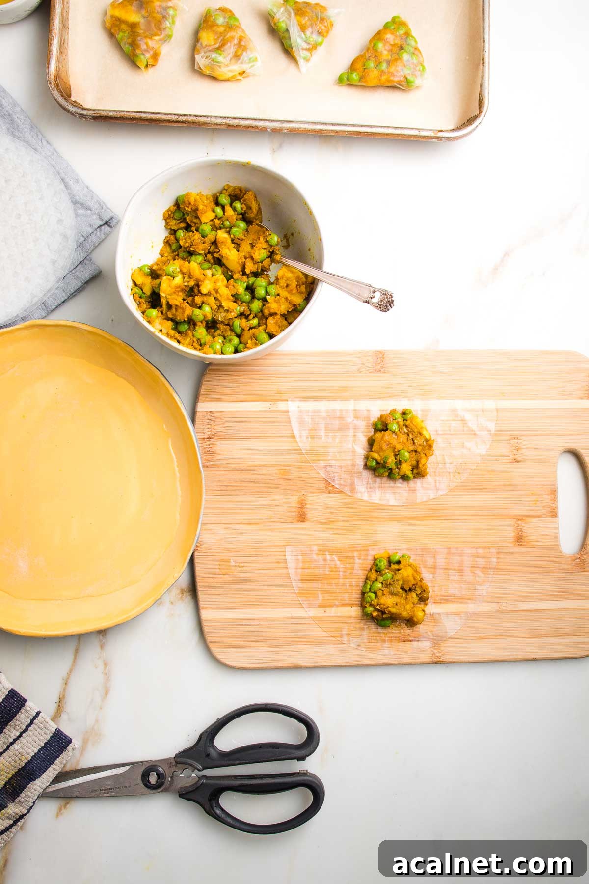 Rimmed baking sheet lined with parchment, a bowl of potato samosa filling, a yellow dish filled with water, a pair of kitchen scissors, and a wooden board with two half moons of rice paper each with a scoop of filling.