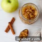 Jar of apple compote next to an apple, cinnamon sticks and a spoonful of compote.
