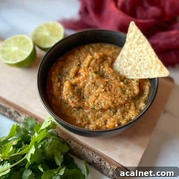 A bowl of pineapple habanero salsa sits on a rustic wooden cutting board surrounded by limes, cilantro, and a red linen.