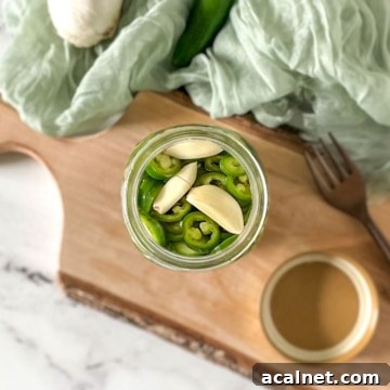 Pickled serrano peppers are shown in a glass jar surrounded by garlic, serrano peppers, and a fork.