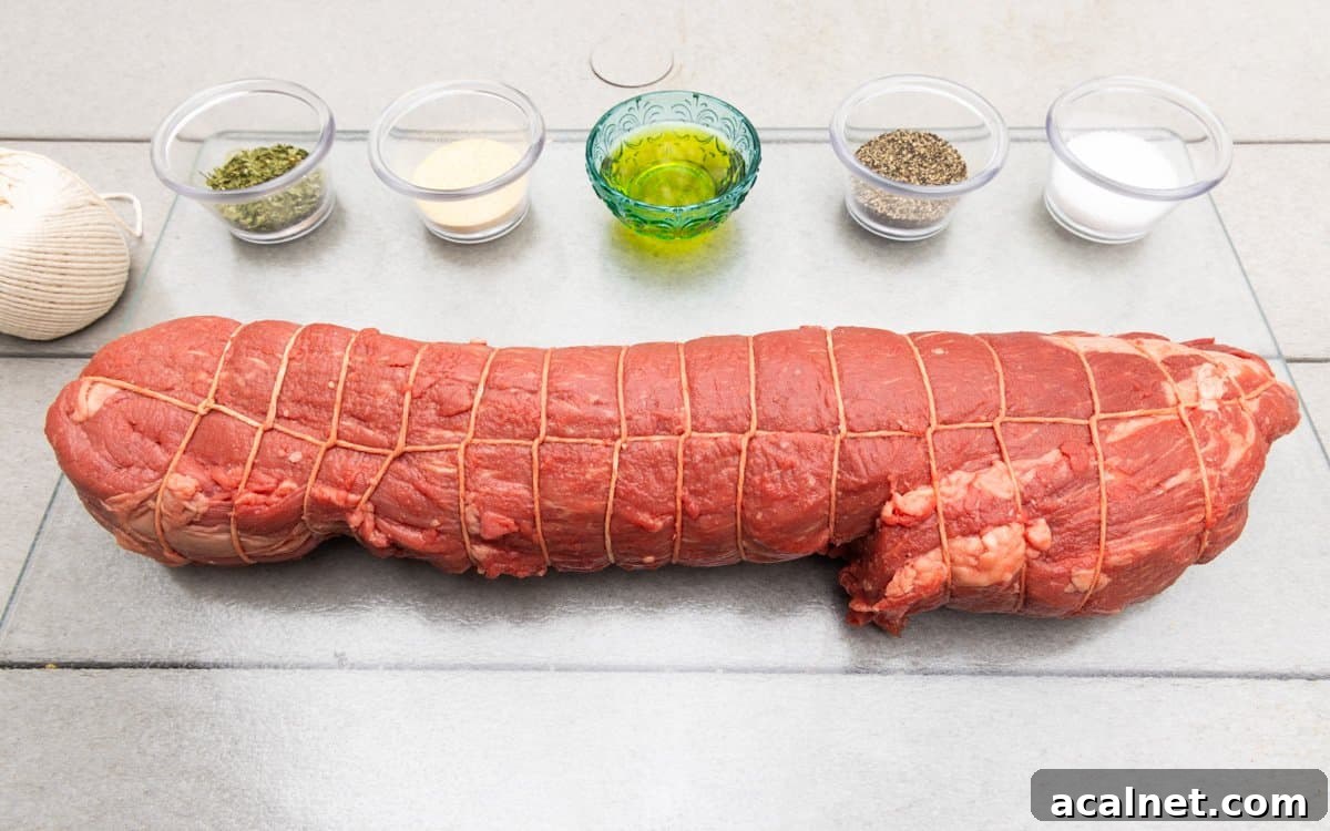Beef tenderloin trussed with butcher string on white wood table with seasonings surrounding the meat in little dishes.