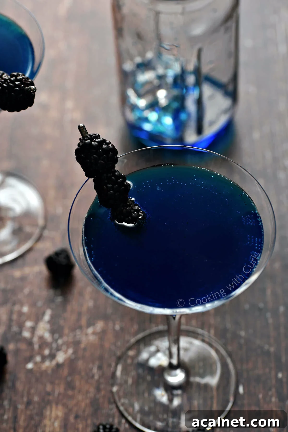 A martini glass filled with a dark royal blue martini that's garnished with three blackberries on a skewer.
