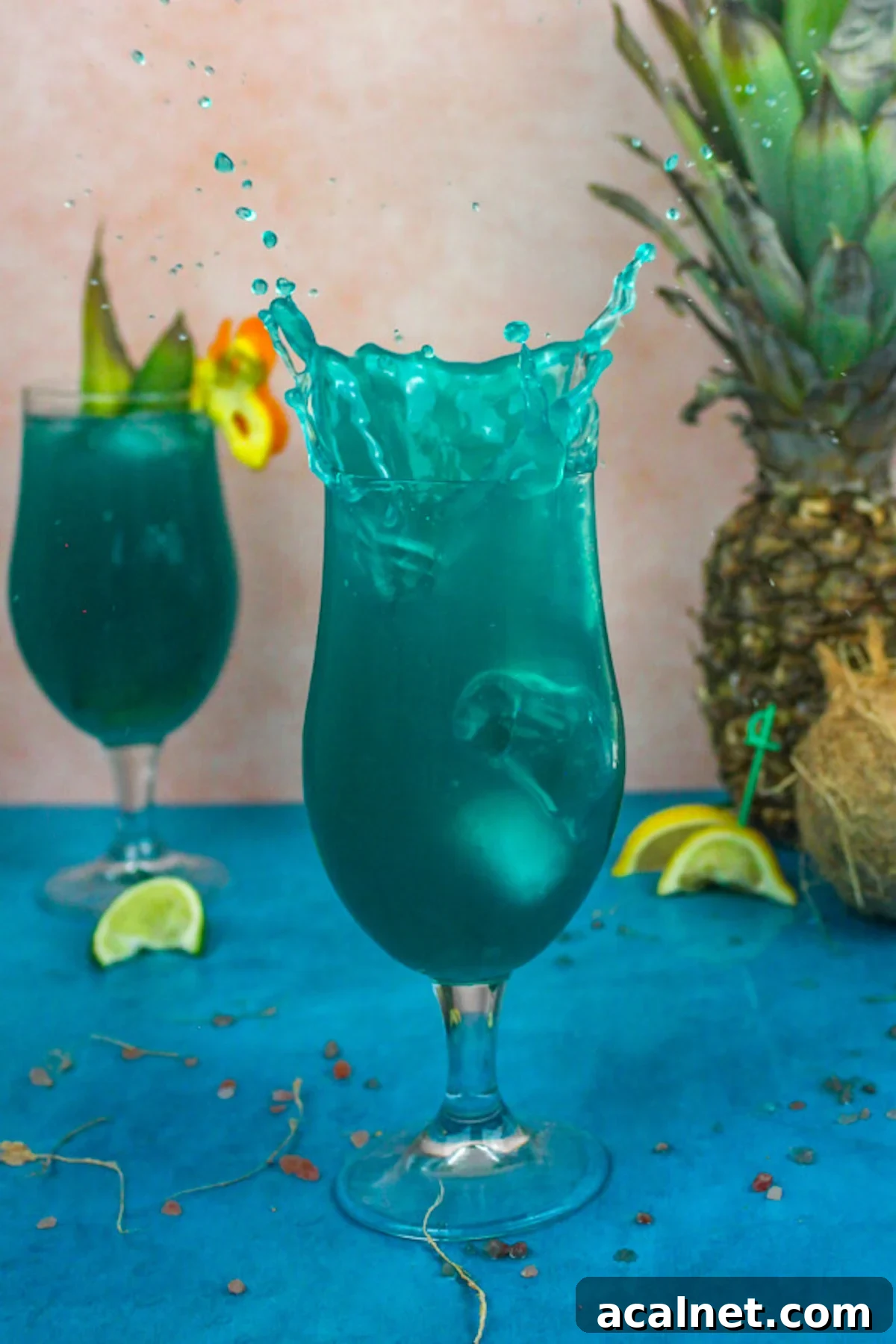 A tall hurricane glass filled with a dark blue pineapple coconut beach cocktail. Ice has been dropped in the drinking and the liquid is splashing out. A pineapple is in the background.