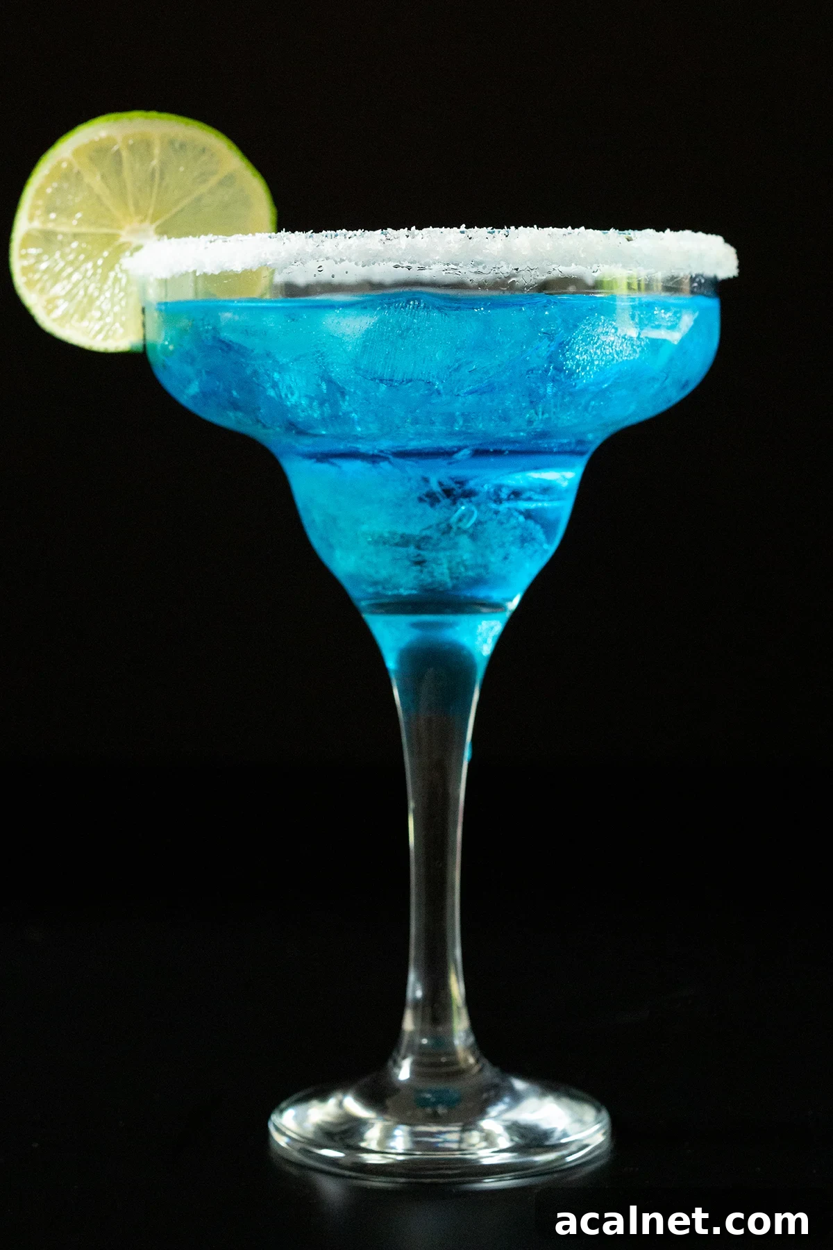 A tall margarita glass is filled with a vibrant bright blue margarita. It's garnished with a salt rim and a lime wheel.