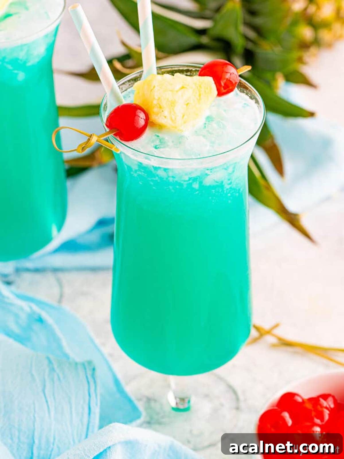 A tall hurricane glass filled with a bright blue Blue Hawaiian cocktail that's garnished with two cherries and a pineapple wedge on a skewer.