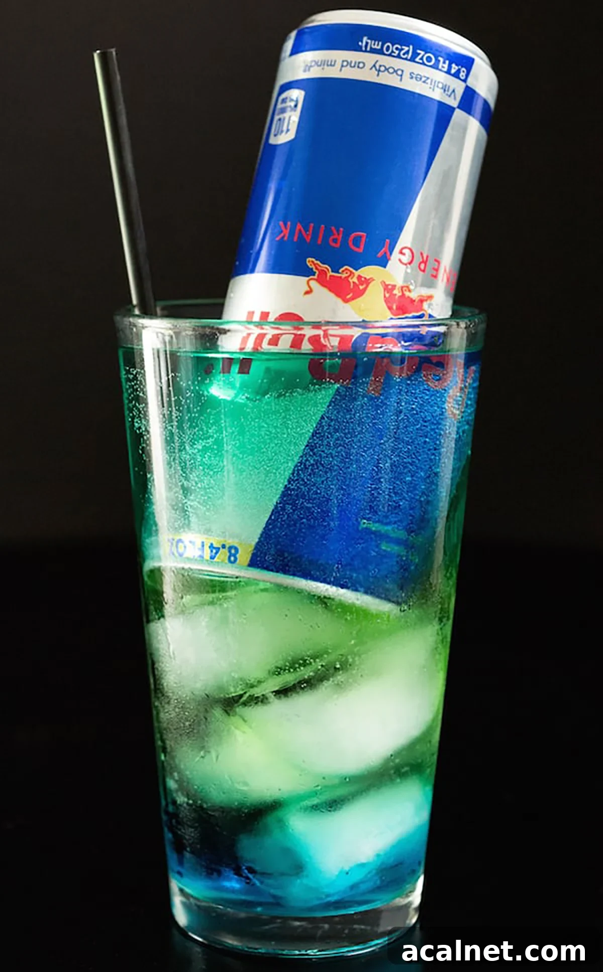 A pint glass has a cocktail that goes from green on the top to blue in the bottom. An entire can of red bull is face down in the glass.