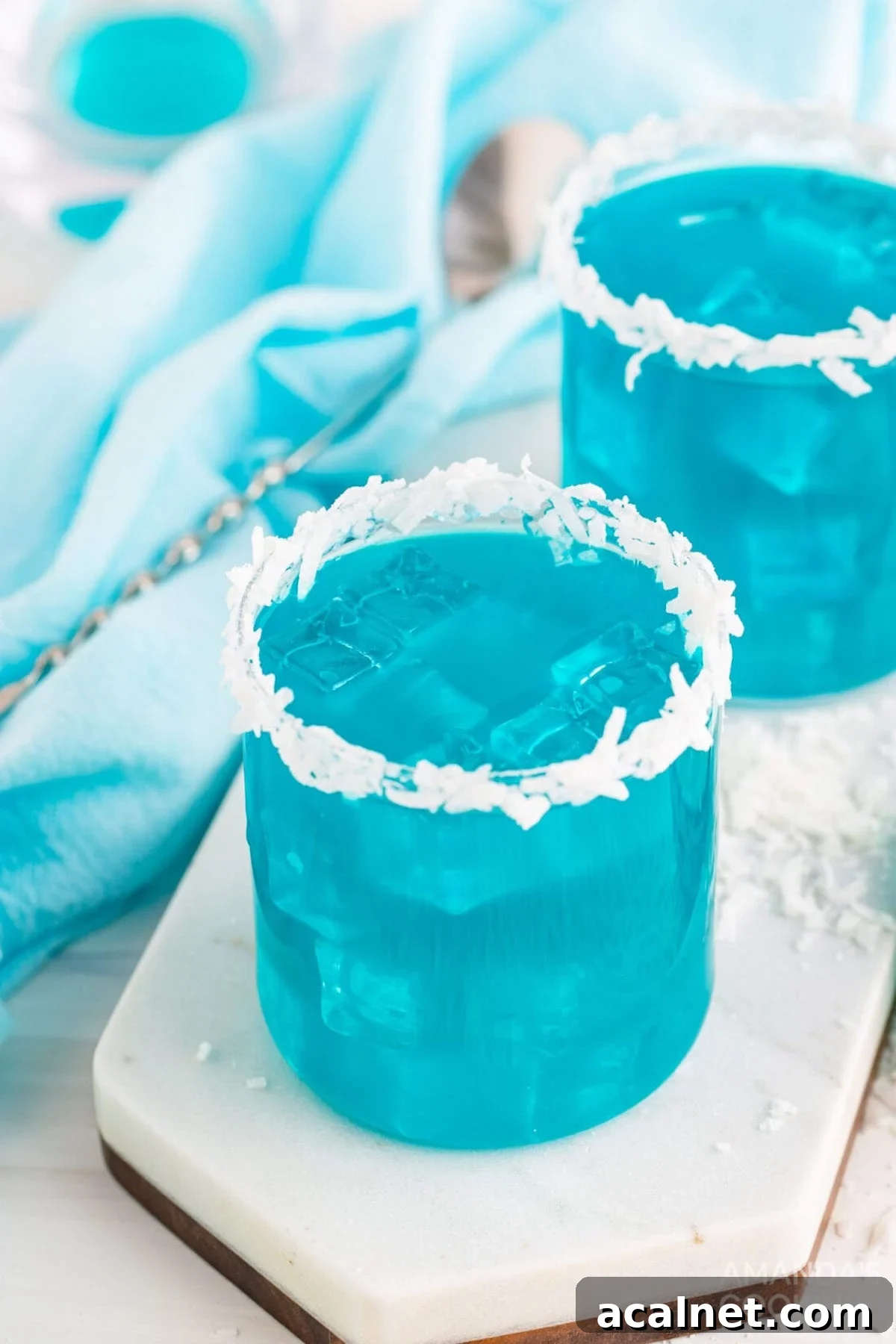 Two old fashioned glasses filled with a bright blue Ocean water cocktail. Each has a rim of shredded coconut.