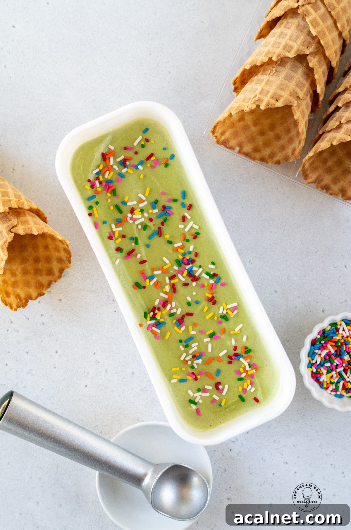 a narrow, long white, rectangular container filled with green matcha ice cream next to rows of waffle cones. Viewed from above