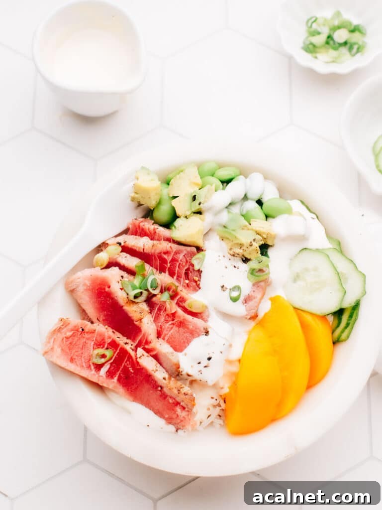 A delicious air-fried tuna steak is served over a bed of rice, topped with dressing, cucumber, edamame beans, and sliced mango. A white spoon rests on the bowl, and small bowls of spring onions are placed nearby.