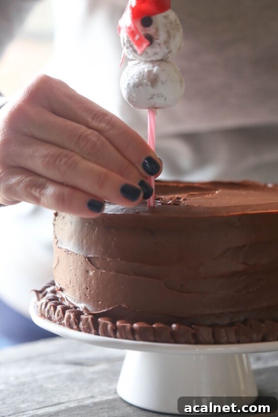 Press the snowmen into the cake. A hand gently pressing the bottom of a cake pop stick topped with a snowman into the top of a frosted chocolate cake.